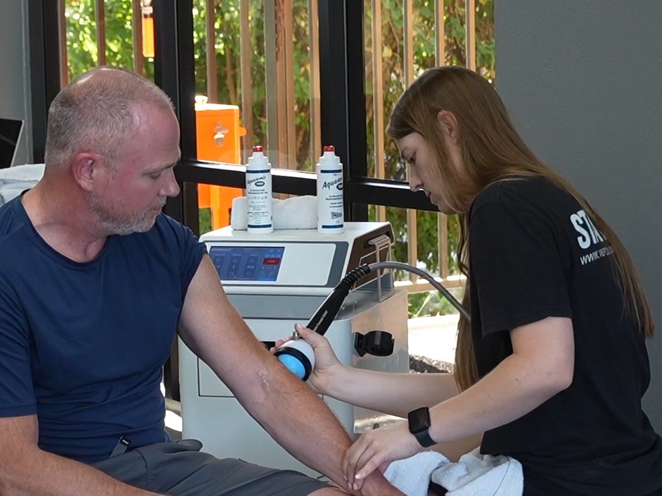A WLF provider pressing the StemWave shockwave handpiece against a seated member's forearm, the console and ultrasound gel behind them.