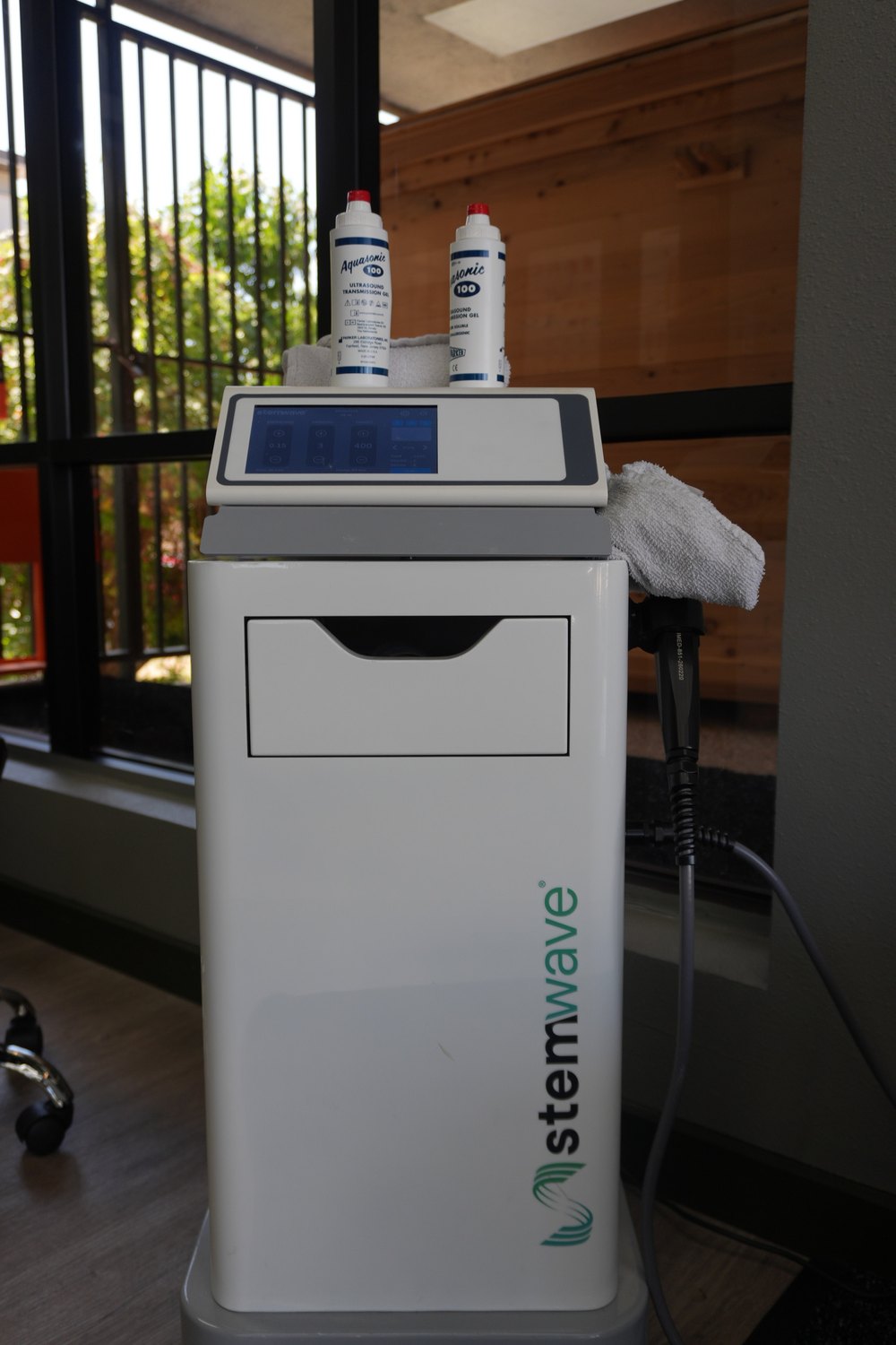 The StemWave shockwave therapy unit at WLF Club: a white console on wheels with its control screen lit, the handpiece coiled on its hose, and ultrasound gel on top, standing by the treatment room window.